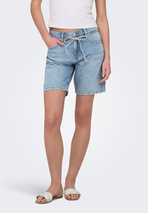 Mujer con pantalones cortos vaqueros azul claro con cordón, camiseta blanca sin mangas y sandalias beige, de pie con una mano en el bolsillo.