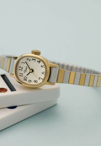 Orologio da polso Timex con cinturino in metallo oro e argento appoggiato su oggetti bianchi impilati su uno sfondo azzurro chiaro.
