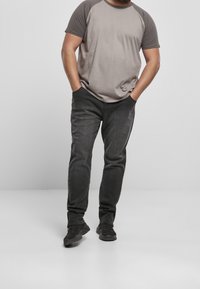 Man klädd i en tvåtonad grå kortärmad skjorta, svarta jeans och svarta sneakers, stående med händerna i fickorna mot en enfärgad bakgrund.