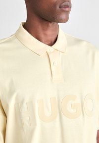 Nahaufnahme einer Person, die ein hellgelbes Polohemd trägt, auf dem große, erhabene Buchstaben "HUGO" über die Brust geschrieben sind.