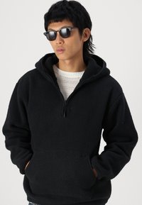 Černý fleecový pulovr s kapucí a polozipem, přední klokankou a texturovanou tkaninou, nošený přes bílý pletený svetr.