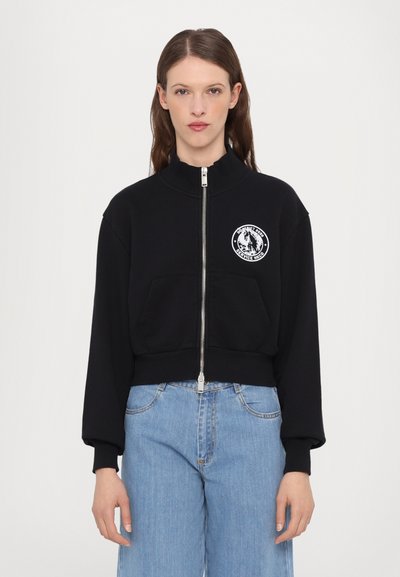Crna skraćena zip-up dukserica s visokim ovratnikom i prednjim džepovima. Sadrži kružni bijeli logo na lijevoj strani prsnog koša. Mekana tekstura.