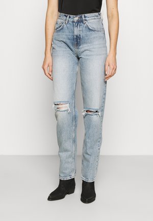 Jeans strappati a vita alta di colore blu chiaro con una vestibilità dritta, caratterizzati da aree rovinate sulle ginocchia e un leggero scolorimento, abbinati a stivaletti neri alla caviglia.