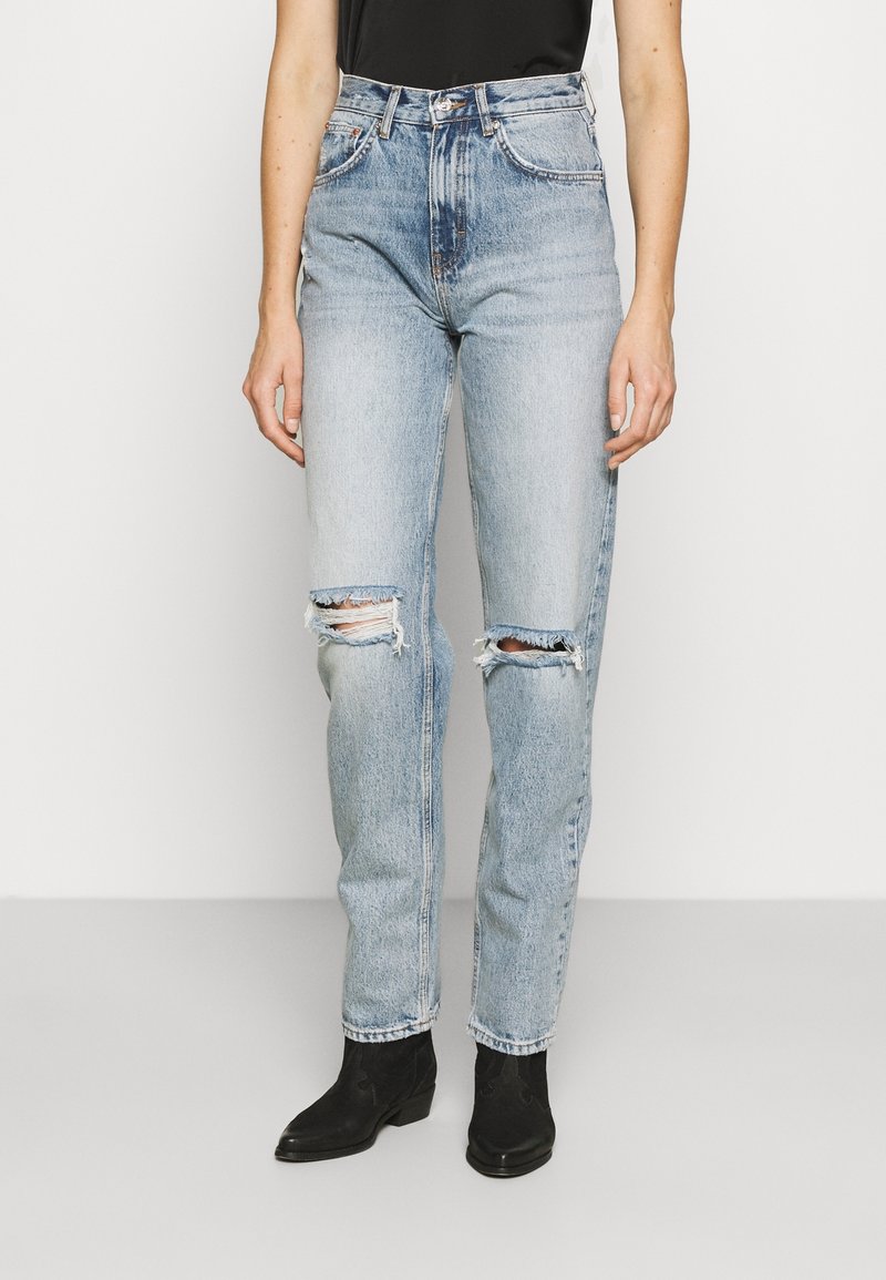 Jeans strappati a vita alta di colore blu chiaro con una vestibilità dritta, caratterizzati da aree rovinate sulle ginocchia e un leggero scolorimento, abbinati a stivaletti neri alla caviglia.