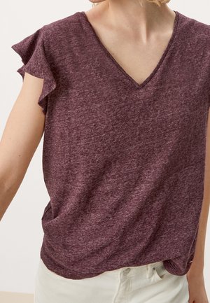 Femme portant un haut à col en V court en jersey bordeaux chiné avec des manches à volants, rentré dans un pantalon blanc cassé.