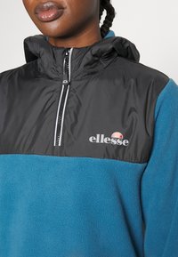 Felpa in pile nera e teal con zip a quarto, dotata di colletto alto, logo sul petto e tessuto testurizzato.