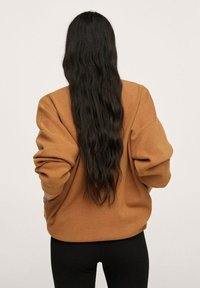 Brązowy, oversize'owy sweatshirt z szerokimi rękawami i teksturowanym materiałem, noszony z czarnymi legginsami. Włosy długie i faliste, opadające na plecy.