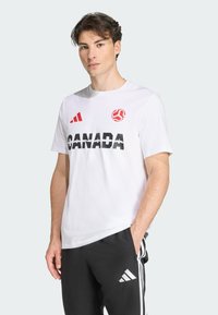 Camiseta blanca de algodón con el destacado texto negro "CANADÁ", acentos rojos y un logo. Combinada con pantalones negros que presentan rayas blancas.