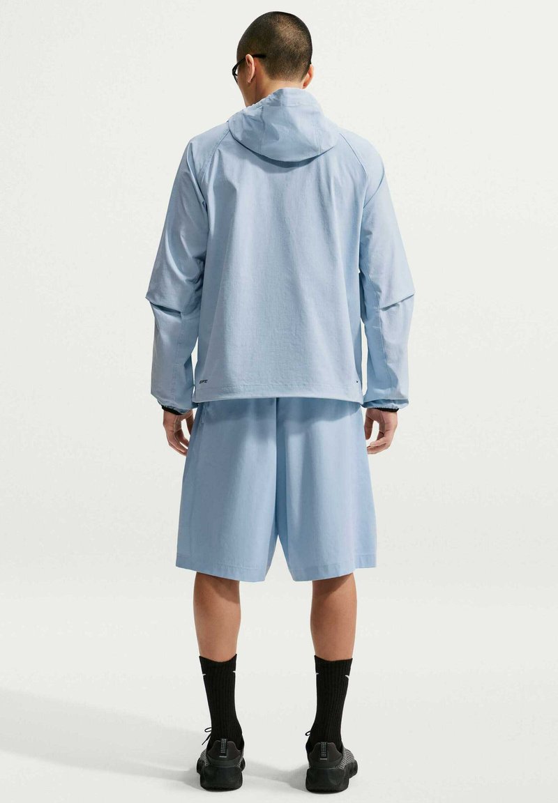 Mann trägt hellblaue Kapuzenjacke und passende Shorts, schwarze Socken und schwarze Sneaker, steht mit dem Rücken zur Kamera vor weißem Hintergrund.
