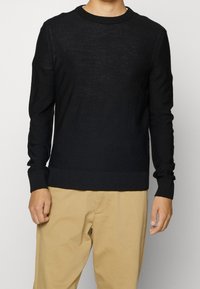 Maglione nero a maglia con collo a giro, polsini e orlo a coste, dotato di un sottile dettaglio del logo. Indossato con pantaloni beige.