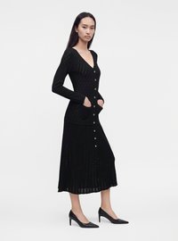 Femme portant une longue robe en maille noire boutonnée avec des poches et des talons noirs, debout sur un fond blanc uni.