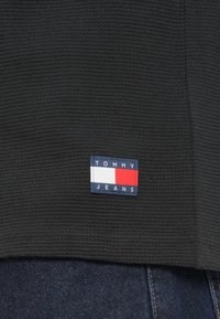 Μαύρο υφασμάτινο ένδυμα με μικρό λογότυπο Tommy Jeans κοντά στο τελείωμα, συνδυασμένο με σκουρόχρωμο μπλε τζιν.