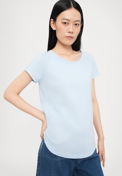 Light blue short-sleeve T-shirt with a scoop neck and curved hem. Fabric appears soft and textured, worn with dark denim jeans.