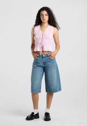 Jonge vrouw met krullend haar die een roze mouwloos gerimpeld topje draagt, denim knielange shorts, witte sokken en zwarte loafers, staand met handen in de zakken.