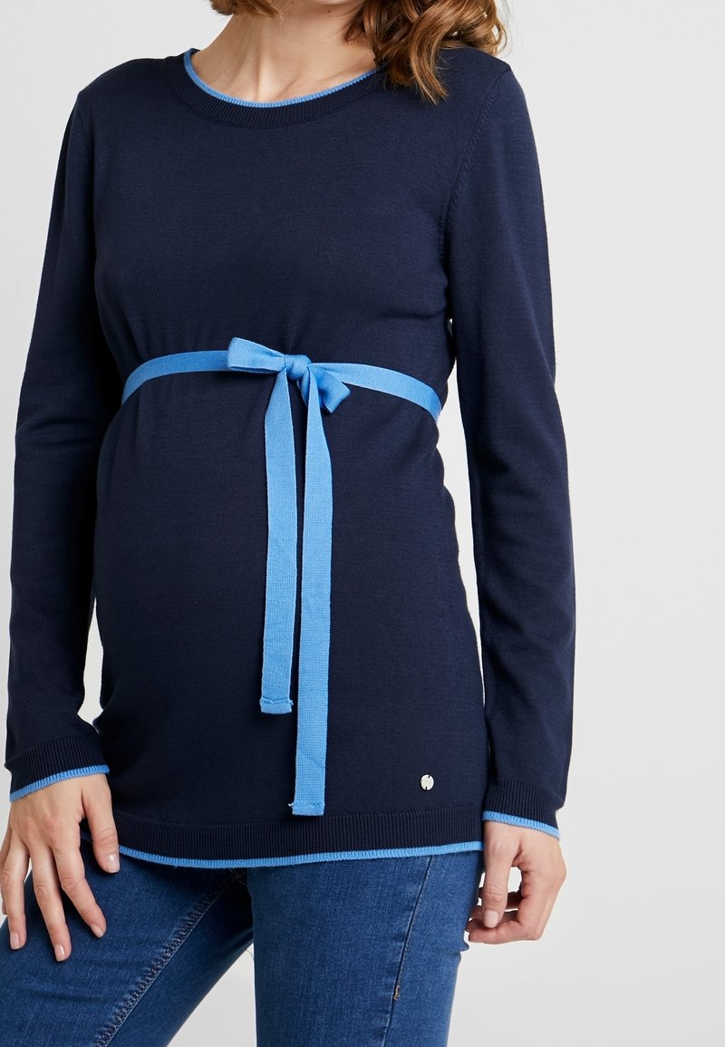 Femme enceinte portant un haut bleu marine à manches longues avec un bord bleu clair et une ceinture nouée en forme de nœud, posant devant un fond uni.