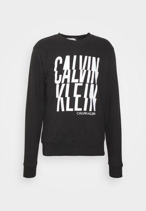 Czarna bluza z długim rękawem z białym, zniekształconym napisem „"CALVIN KLEIN"” na przodzie i mniejszą nazwą marki po prawej stronie u dołu.