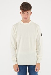 Pullover beige chiaro con scollo rotondo, maniche lunghe, polsini a costine e una toppa triangolare nera sulla manica sinistra. Texture morbida.