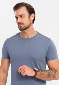 Harmaa lyhythihainen t-paita pehmeästä kankaasta klassisella pyöreällä kaula-aukolla; siinä on hienovarainen tekstuuri, ja se on yhdistetty ruskeaan kelloon ja käsivarren tatuointiin.