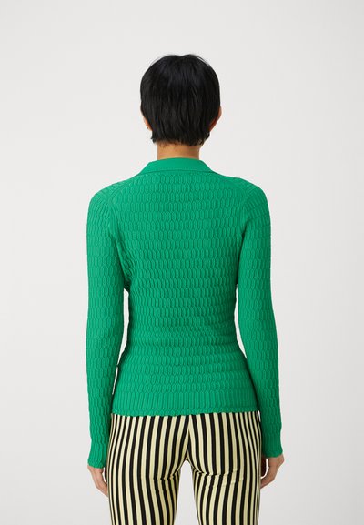 Top tricotat ajustat, verde, cu un model texturat, manșete lungi și guler, asortat cu pantaloni în dungi negre și albe.