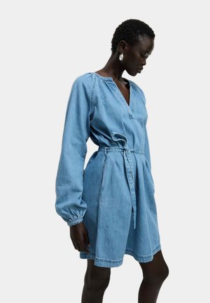 Vrouw met kort haar draagt een lichtblauwe denimjurk met ceintuur, lange opgeblazen mouwen en zilveren hoepeloorbellen, kijkt naar beneden.