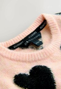 Rozā adīta džemperis ar apaļu kakla izgriezumu, kas rotāts ar lielu melnu sirds dizainu. Audums ir mīksts un pūkains; uz etiķetes ir norādīts zīmola nosaukums "AllSaints."