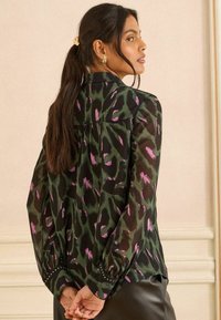 Caurskrāpums zaļā krāsā ar abstraktu melnu un rozā rakstu, garām pūkainām piedurknēm un rotātiem piedurkņu galiem. Brīva piegriezuma ar aizmugures kroku.