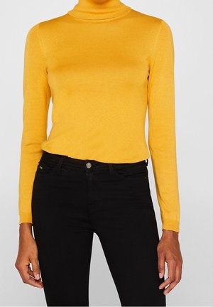 Żółty sweter z golfem o długich rękawach, o prążkowanej fakturze i dopasowanym kroju, zestawiony z czarnymi dżinsami z przodem zapinanym na guziki.