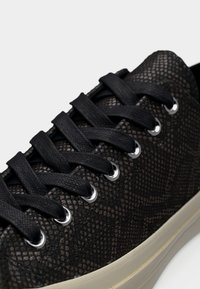 Μαύρο αθλητικό παπούτσι με υφή που θυμίζει δέρμα φιδιού, μαύρα κορδόνια περασμένα μέσα από ασημένιες οπές και παχύ λευκό πάτο.