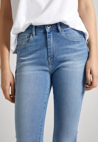 Jeans de denim azul claro con un diseño ajustado, que cuentan con bolsillos estándar y un cierre de botón. Textura suave con un leve desgaste.
