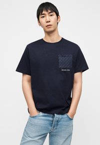 Μπλε βαμβακερό t-shirt με τετράγωνη τσέπη που διαθέτει εκτύπωση λογοτύπου MK σε τονικά χρώματα, στρογγυλή λαιμόκοψη και άνετη γραμμή. Φορεμένο με ανοιχτόχρωμα τζιν.