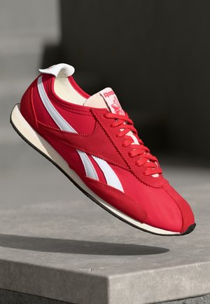 Punane sportlik jalats valgete triipudega, nubuk- ja võrgumaterjal, lame kummist tallaga, ümar varvas ja pehme tekstiilist keel.