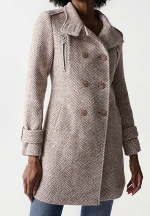 Kvinde iført en beige sildebensmønster uldfrakke med dobbeltradede knapper, lynlåslommer på brystet, manchetstropper og blå jeans.