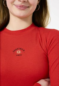 Maglietta rossa a maniche lunghe con collo alto, con testo nero che recita "INSIEME" e "GARICA," accentuata da una grafica di sole.