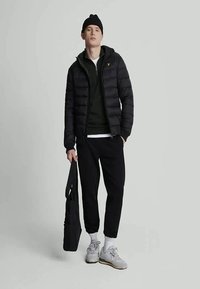 Ung man i svart beanie, svart pufferjacka, svarta joggers och vita sneakers, håller en svart totebag mot en enfärgad bakgrund.