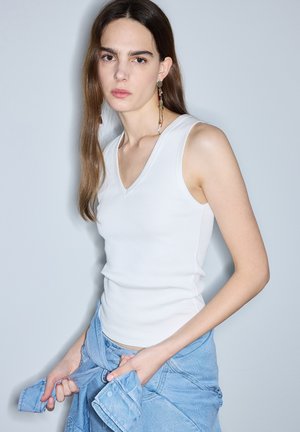 Vrouw met lang bruin haar draagt een wit mouwloos topje en hangende oorbellen, houdt een blauwe spijkerjas vast die om haar taille is gestrikt.