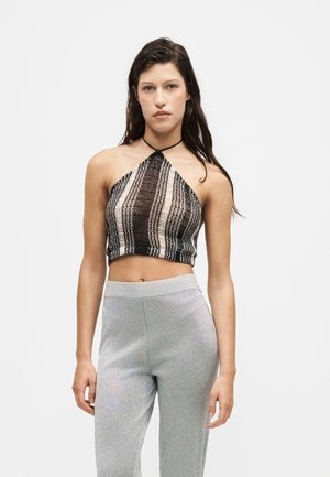 Jeune femme aux longs cheveux foncés, portant un crop top halter rayé noir et blanc et un pantalon argenté taille haute sur un fond uni.