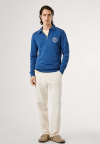 Mėlynas ketvirčio užtrauktukas džemperis su gyslotais rankoviciais, priekinėmis kišenėmis ir logotipo detale, derinamas su grietinėlės spalvos kelnėmis ir rudos spalvos slydimo bateliais.