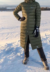 Langer olivgrüner Steppmantel mit durchgehendem Reißverschluss und Seitentaschen, kombiniert mit schwarzer Hose und grauen gefütterten Stiefeln auf verschneitem Gelände.
