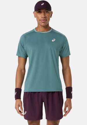 Man met een blauwgroen sportshirt, bordeauxrode shorts, bijpassende polsbandjes en een bordeauxrode pet, staande tegen een effen achtergrond.