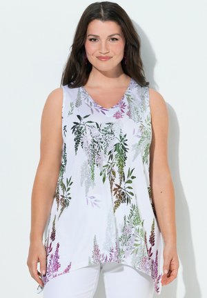 Kvinde iført ærmeløs hvid top med flerfarvede blomsterprint, kombineret med hvide bukser, stående mod en ensfarvet baggrund.