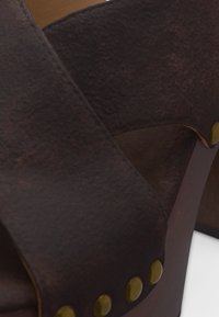 Κοντινή λήψη ρέλι καφέ δέρματος σαγιονάρων με ορείχαλκινες διακοσμήσεις σε ξύλινη σόλα σε απλό φόντο.