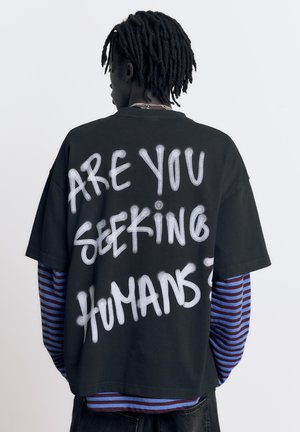 Person med dreadlocks iført en sort T-shirt med hvid tekst "SØGER DU MENNESKER?" over en langærmet trøje med blå og mørkerøde striber.