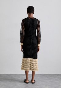 Donna in piedi con la schiena verso la telecamera, indossa un vestito in rete nero con frange in beige a strati sui polsini e sull'orlo, scarpe nere senza lacci, di fronte a un muro grigio.