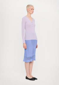 Lavendelkleurig langslavend shirt met knopen, gecombineerd met een lichtblauwe knie-lange rok met een ruchesrand en doorschijnende afwerking. Zwarte flats maken de look af.
