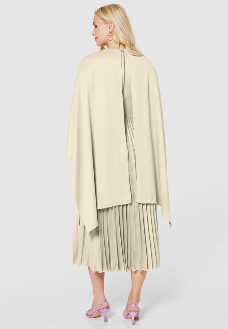 Lichtbeige cape boven een geplooide jurk, met zacht stof en een hoge hals met een sluiting aan de achterkant. Accentdetails zijn lavendelkleurige hakken.