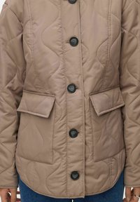 Bež prešita jakna z valovitim vzorcem, črnimi gumbi in dvema sprednjima žepoma. Tkanina je gladka in rahlo sijajna.