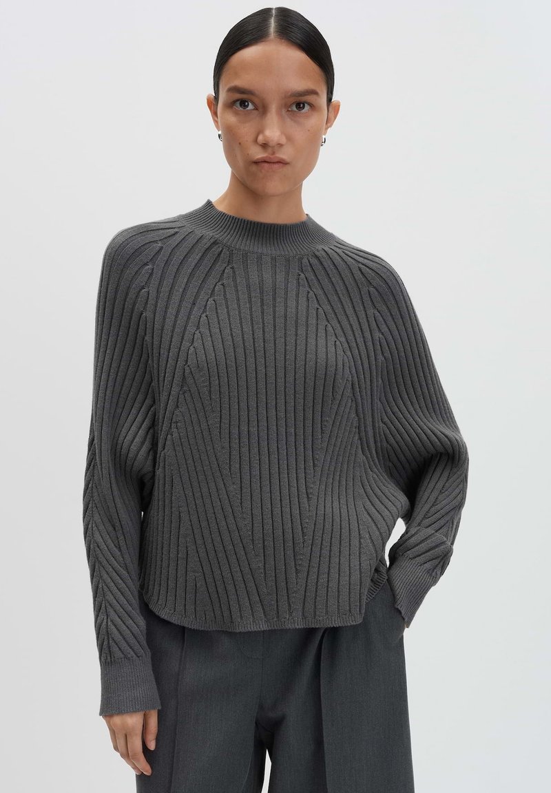 Donna con i capelli raccolti, che indossa un maglione a costine grigio scuro e pantaloni a gamba larga abbinati, in piedi davanti a uno sfondo chiaro e semplice.