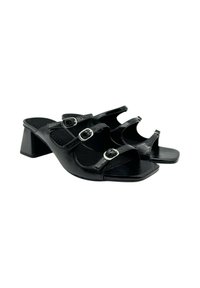 Sandalias de tacón negro con tres correas ajustables, hebillas de plata, punta descubierta y tacón bajo. Textura de cuero suave y suela plana.