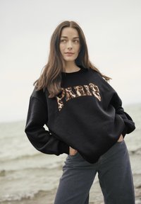 Felpa oversize nera con la scritta "PARIS" in lettere leopardate, abbinata a jeans grigio scuro, il tutto in un contesto costiero.