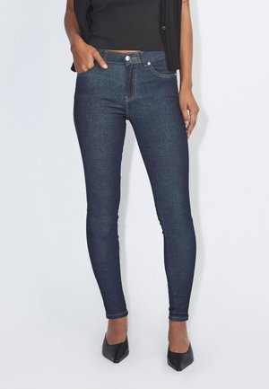 Mujer con vaqueros pitillo azul oscuro con bolsillos delanteros, zapatos negros de punta puntiaguda y un top negro corto sobre un fondo blanco.